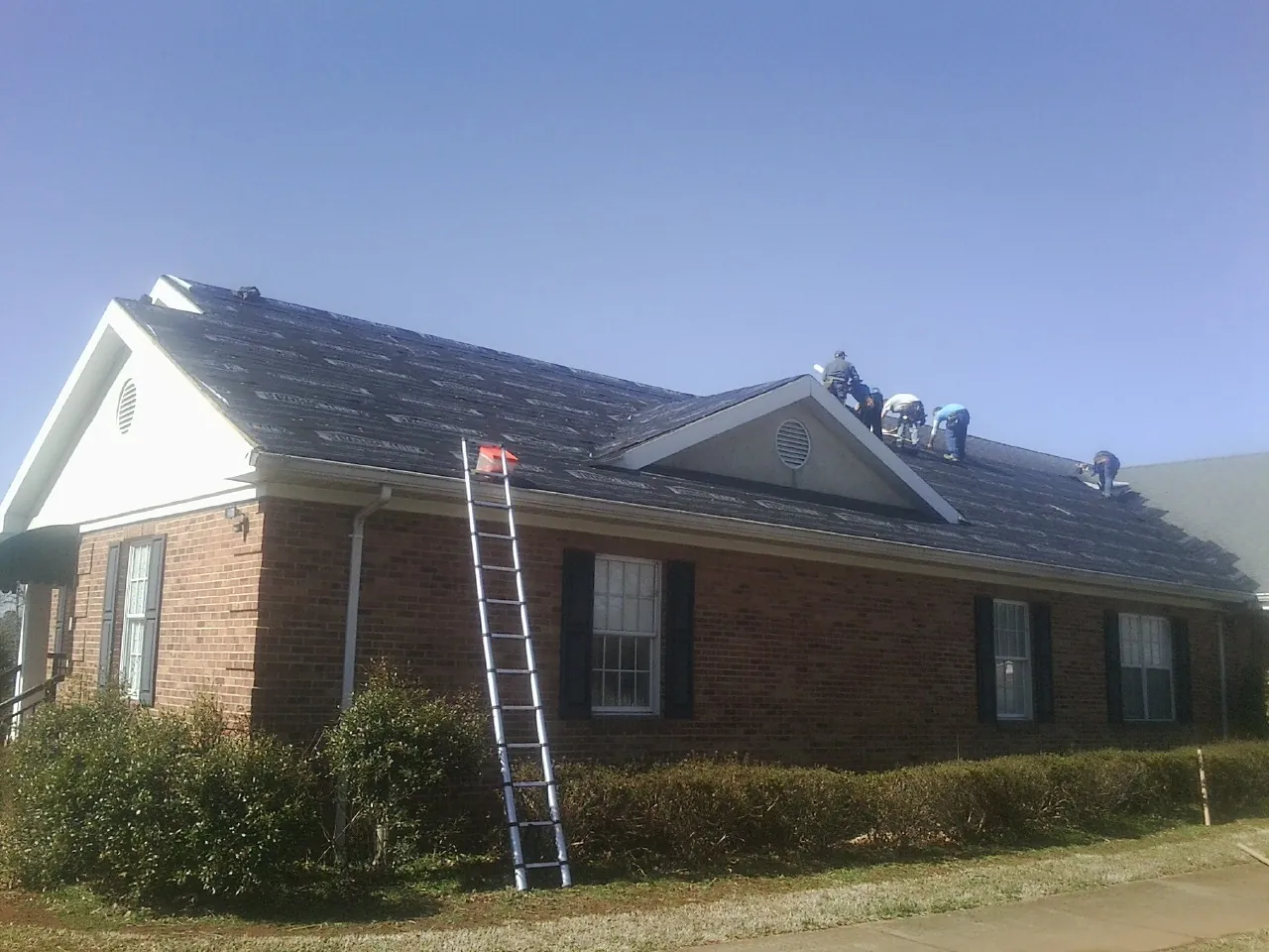 Crew tearing shingles off a brick ranch roof with a ladder set — Emergency Roof Repair work by our crew in Justice