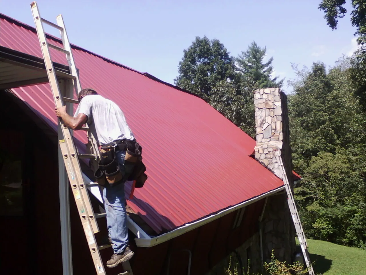 Roofer on a ladder beside a red metal roof and stone chimney — Soffit & Fascia Repair by Roofing Brothers in Justice